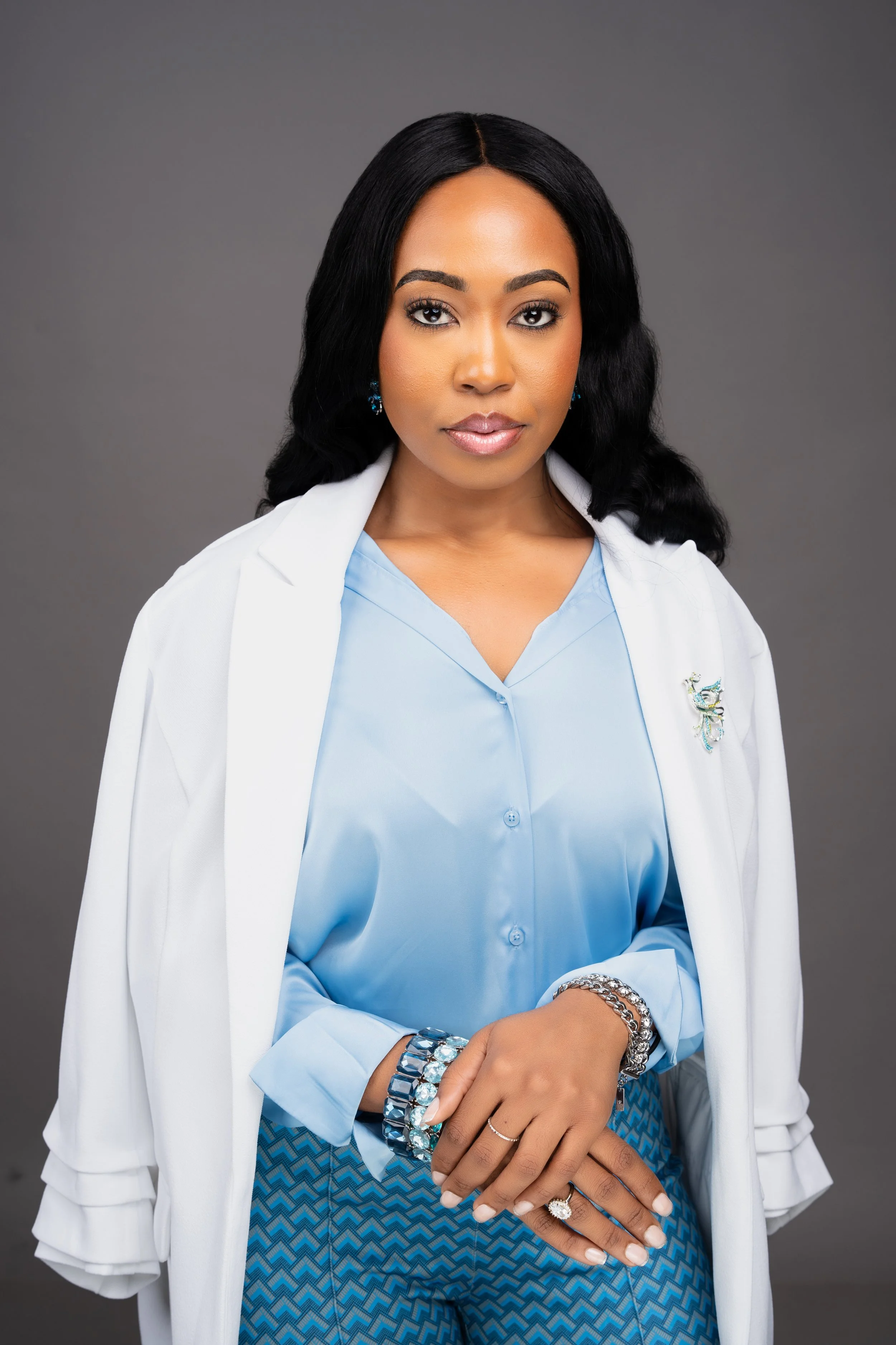 A woman with black hair, brown skin, and makeup, wearing a white blazer and a light blue blouse with jewelry, standing against a gray background.