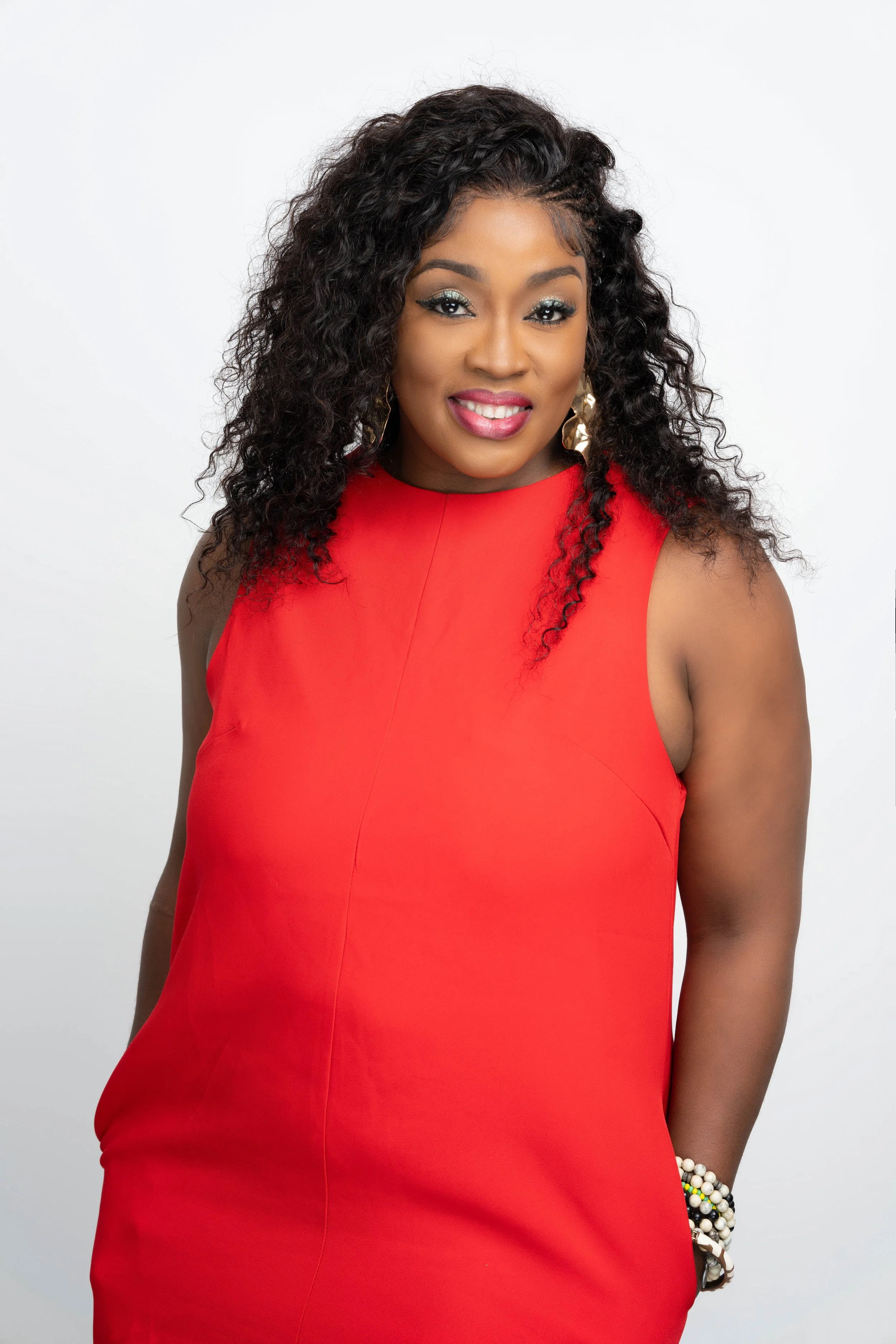 A woman with curly black hair, wearing a red sleeveless dress, smiling with makeup and earrings, standing against a white background.