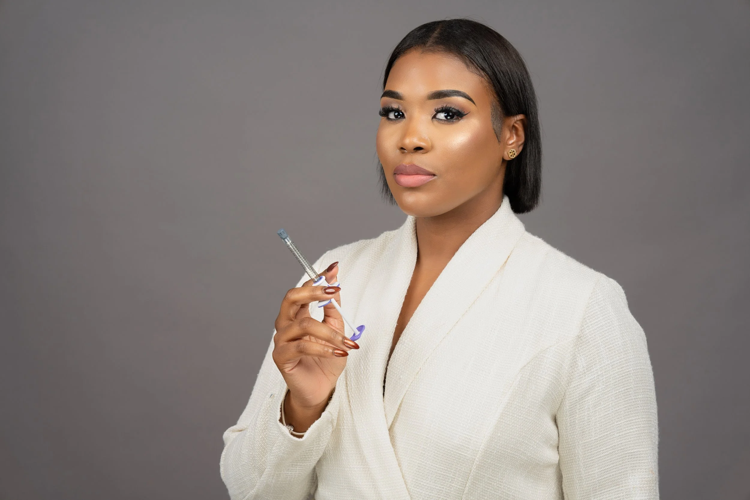 A woman with dark hair and makeup, wearing a white blazer, holding a syringe, standing against a gray background.
