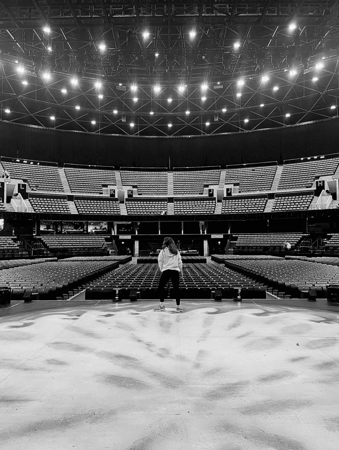 A person standing on a stage in an empty theater or concert hall with multiple levels of seating, overhead stage lighting, and a large open space.