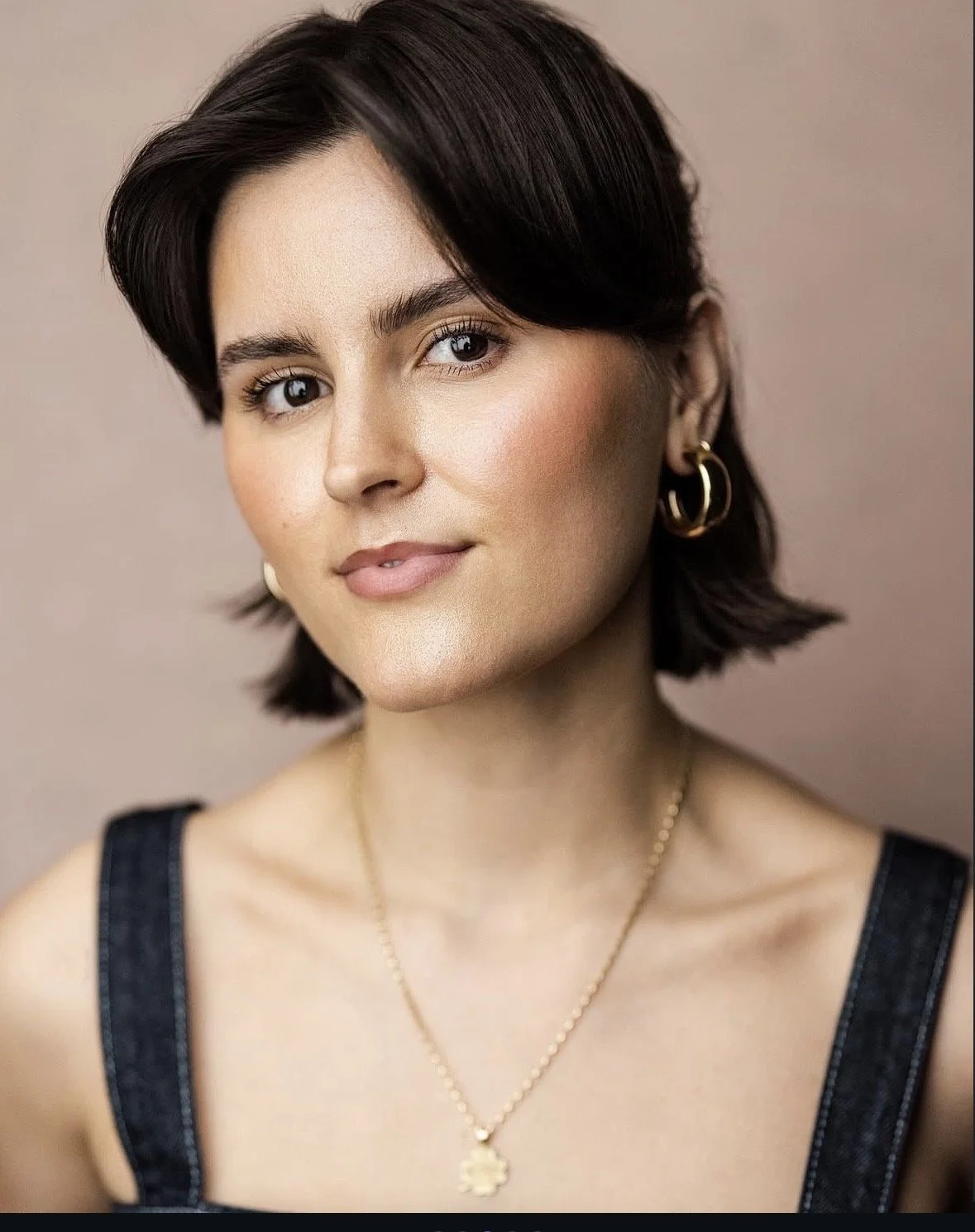 A young woman with short dark hair and light makeup, wearing gold hoop earrings, a gold necklace, and a black sleeveless top, looking at the camera with a slight smile.
