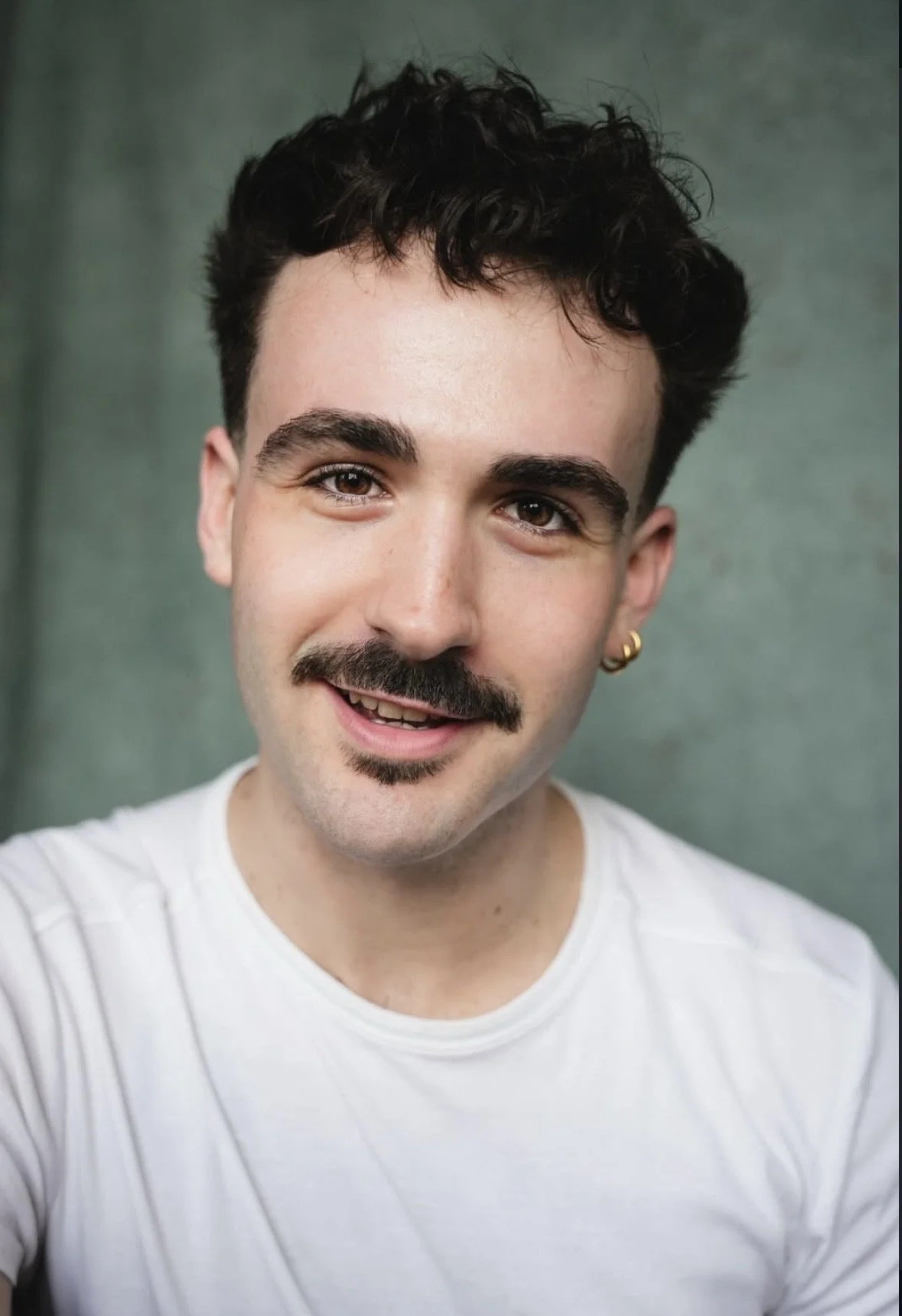 A young man with dark, curly hair, a mustache, and a small beard, wearing a white T-shirt and a gold earring in his left ear, smiling at the camera against a greenish background.