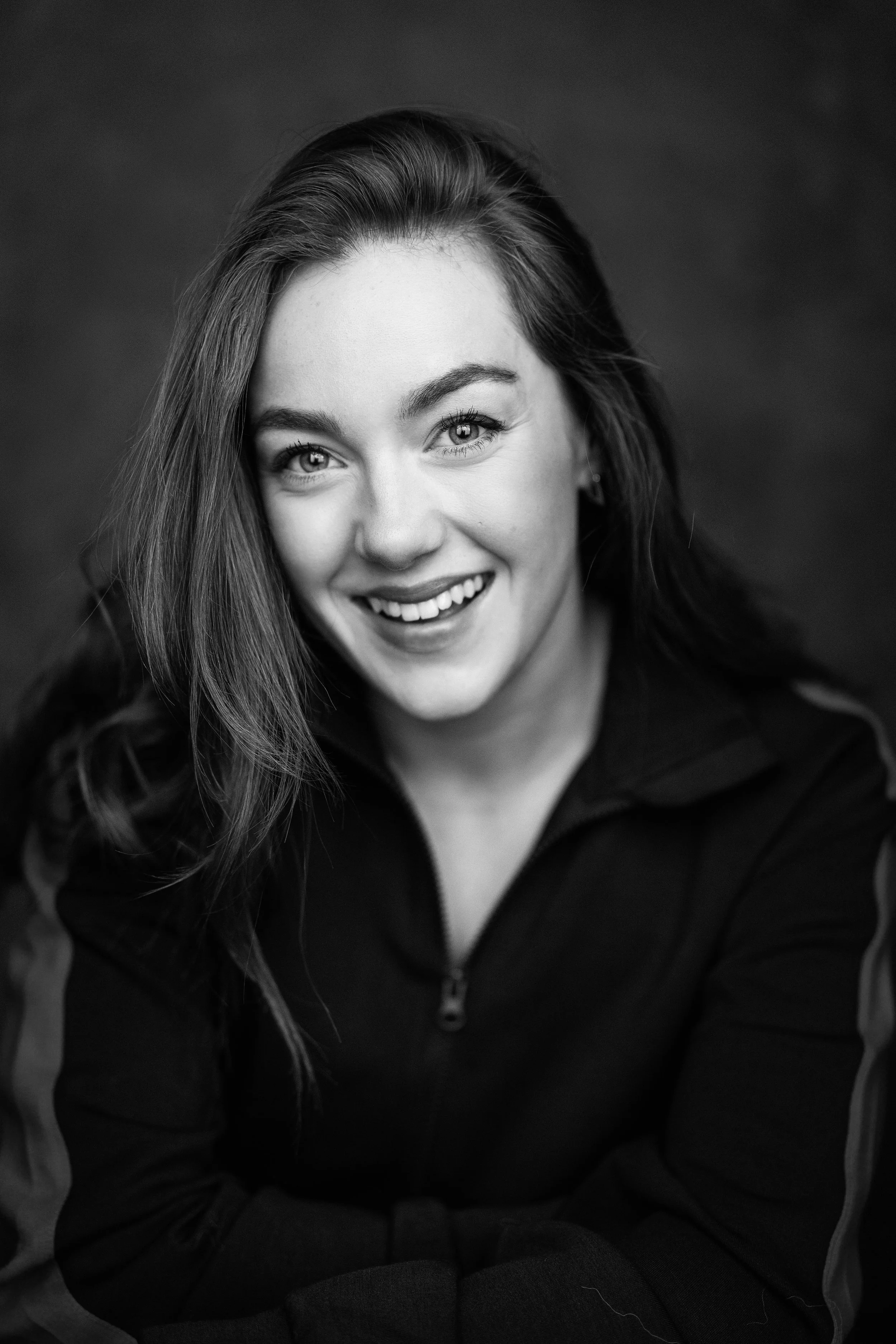Black and white portrait of a smiling young woman with long hair, wearing a jacket with a zip, looking at the camera.