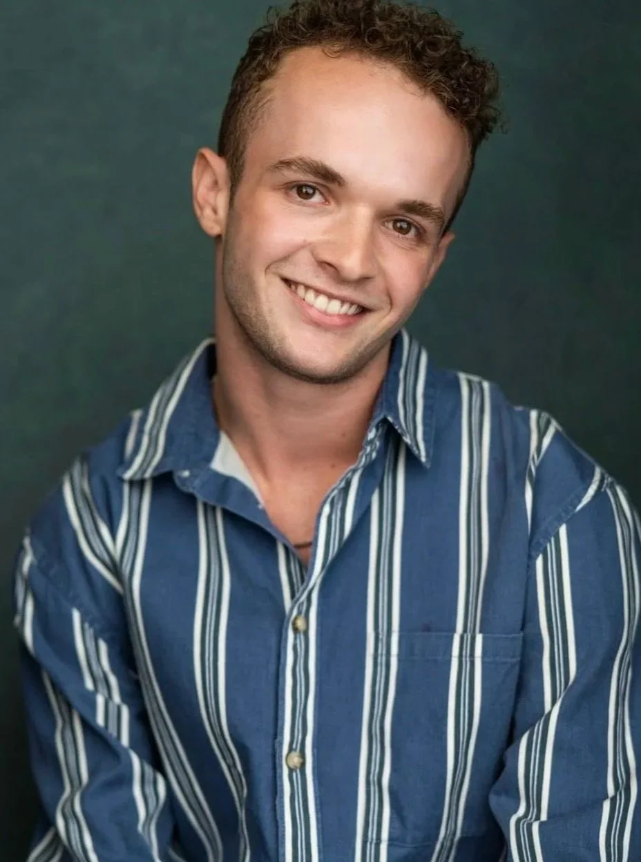 A young man with short curly hair smiling, wearing a blue and white striped button-up shirt, against a dark green background.