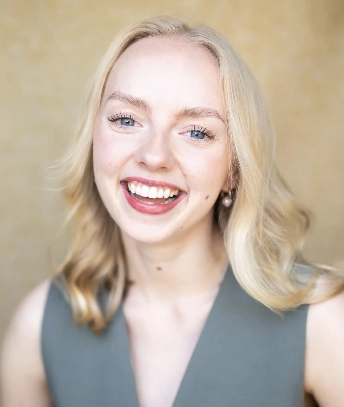 A young woman with blonde hair, blue eyes, and a bright smile, wearing earrings and a sleeveless gray top, against a warm background.