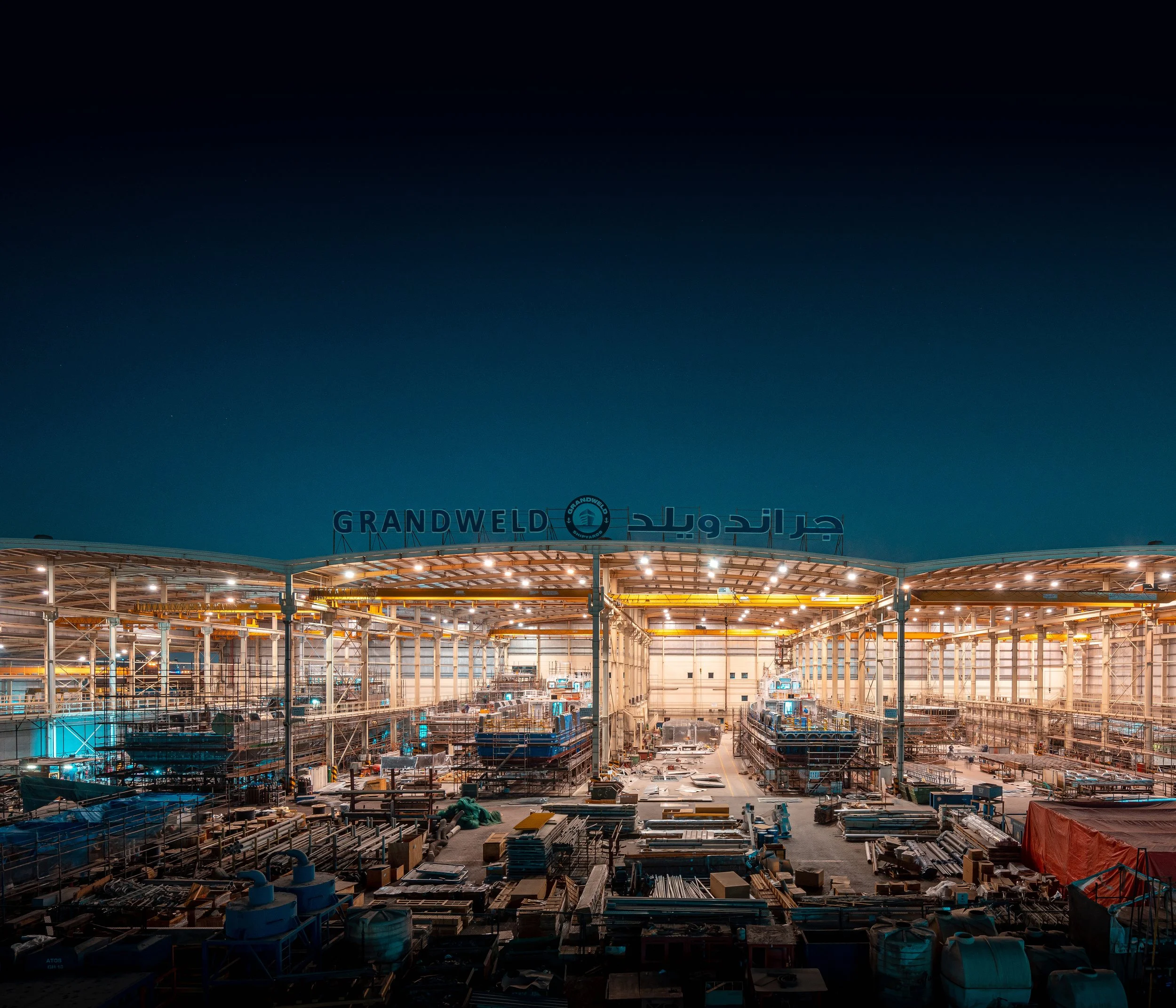 Construction site at night with scaffolding, building materials, and equipment under bright lights, with a sign displaying 'GrandWeld' and Arabic script.