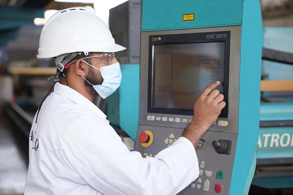 A factory worker wearing a white hard hat, face mask, and safety glasses operating a large industrial machine with a touchscreen interface.