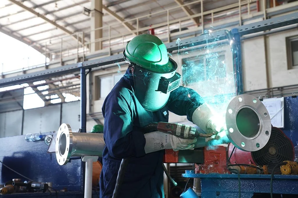 A worker wearing a green welding helmet and protective gloves welding a metal pipe in an industrial setting.