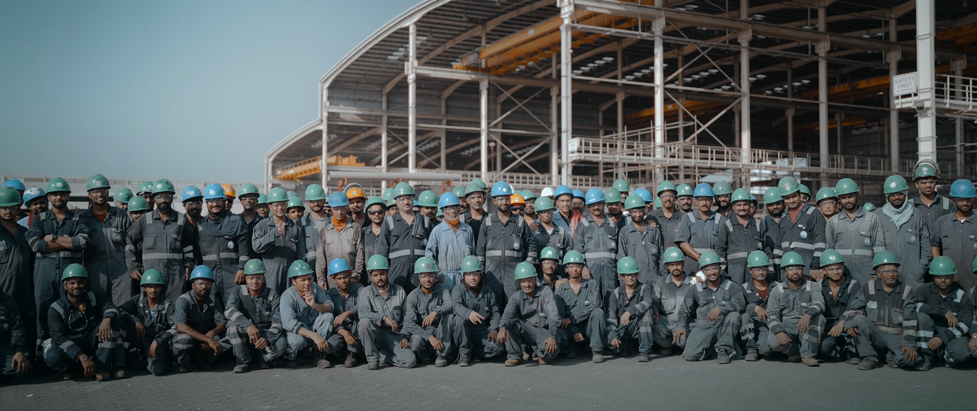 Group of construction workers wearing safety helmets and work uniforms in front of an industrial building under construction.