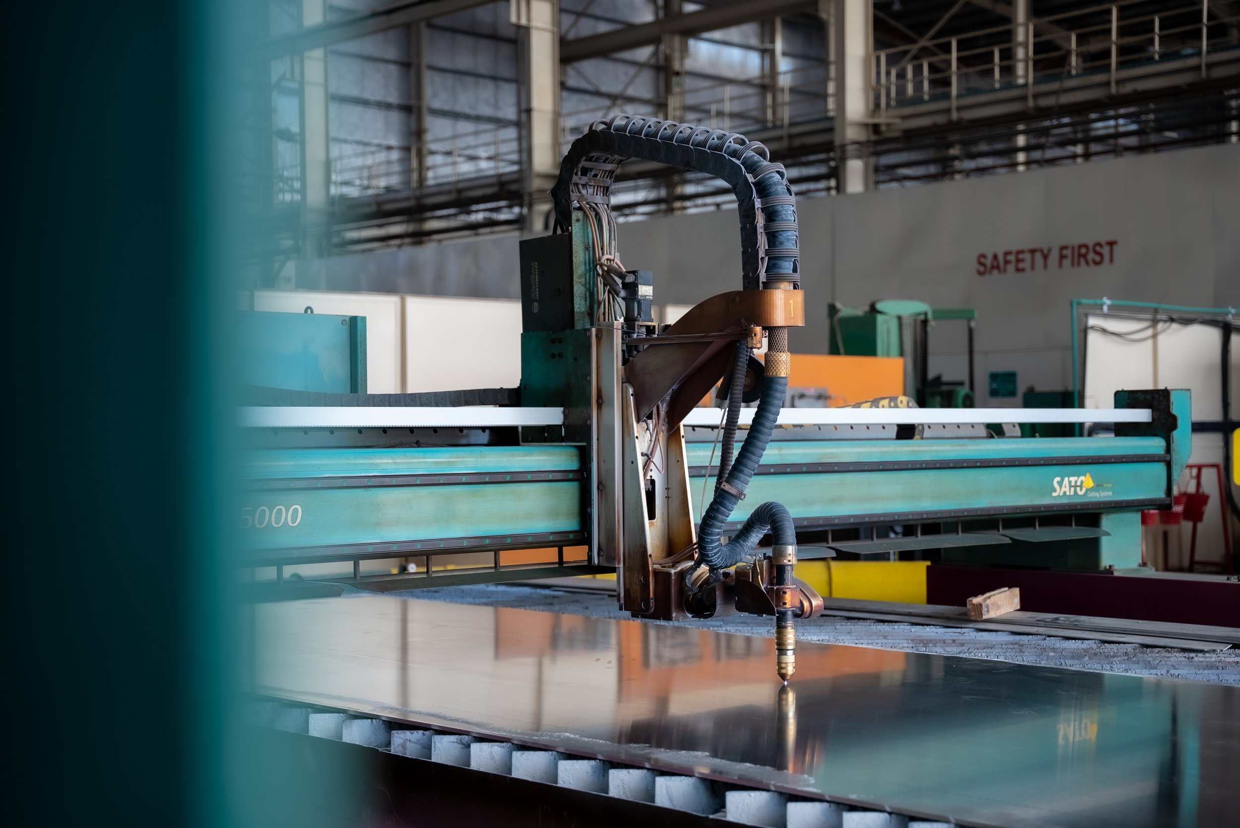 Industrial machine in a factory setting, with a robotic arm and various cables, and safety signage reading 'SAFETY FIRST' in the background.