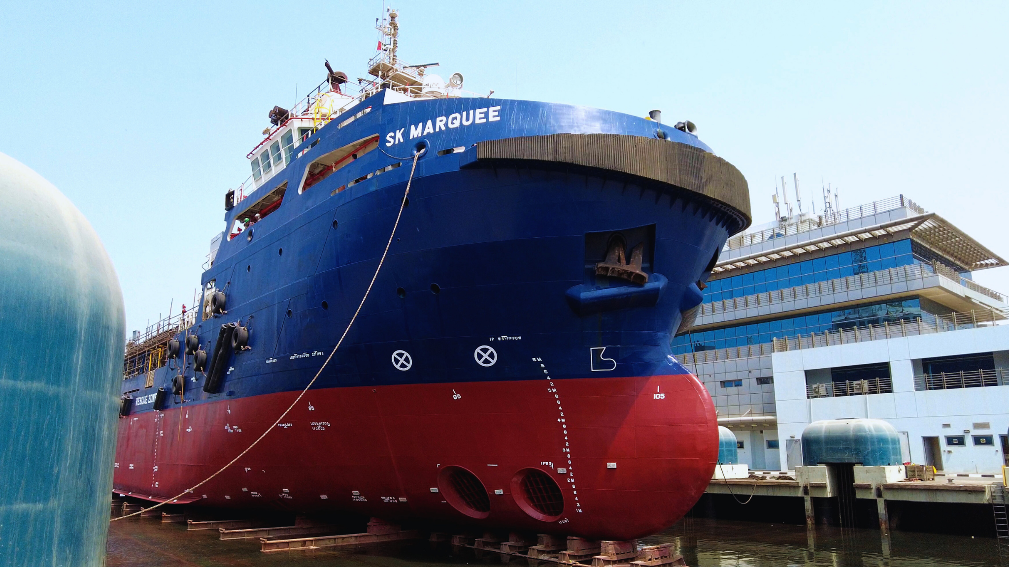 A large blue and red ship named SK Marquee docked at a port with modern buildings in the background.