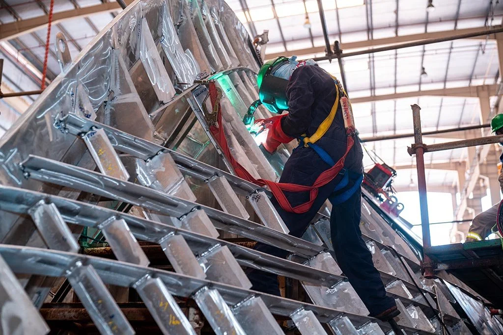 A worker welding a large metal structure inside an industrial workshop, wearing safety gear including a helmet and harness.