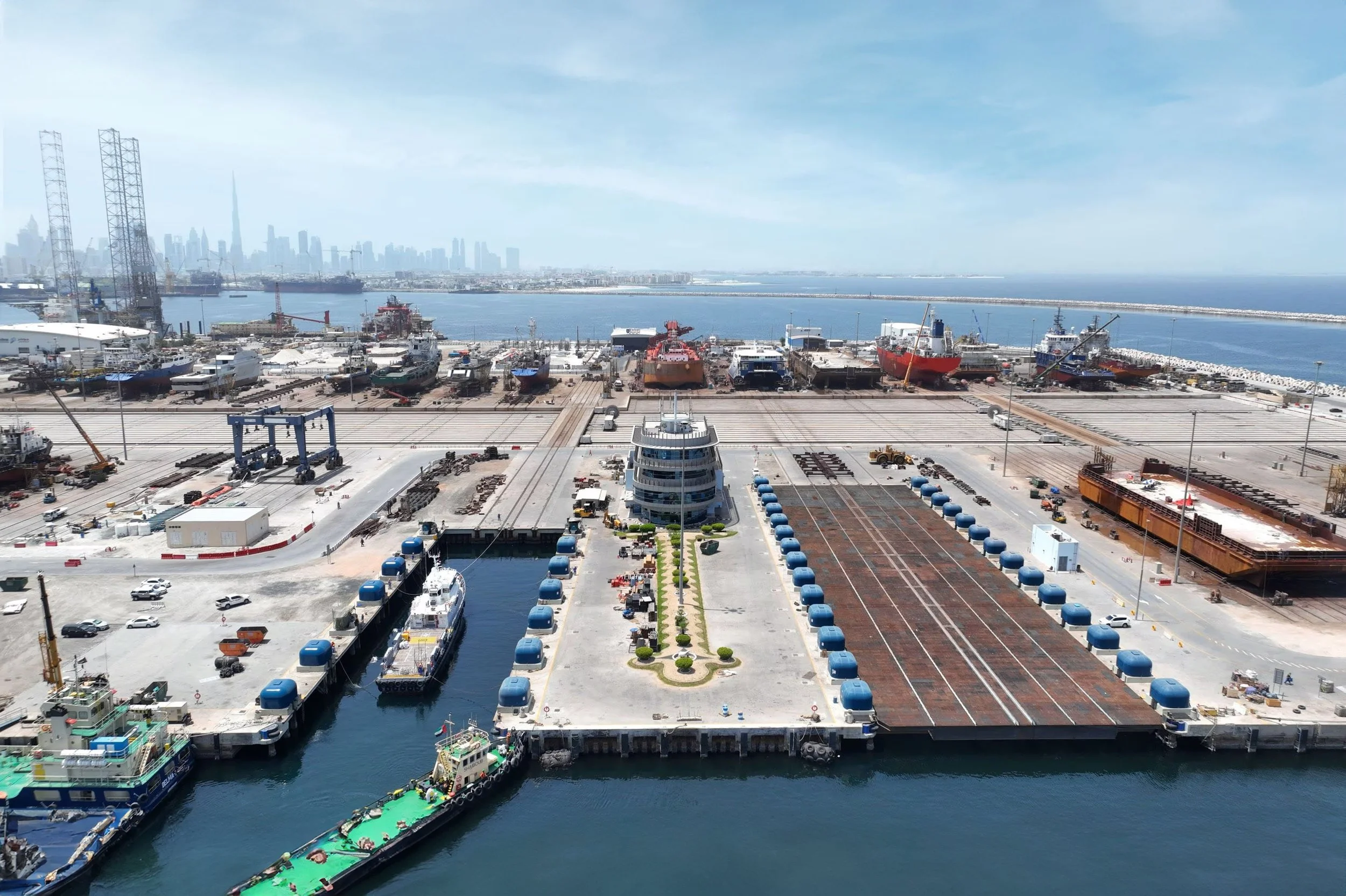 A busy port with boats and ships docked, a waterfront city skyline in the background, and a clear blue sky.