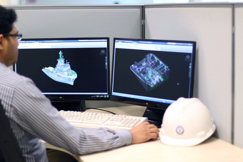 A person working at a desk with dual monitors displaying 3D models of a ship and a building, with a white hard hat on the desk next to the keyboard.