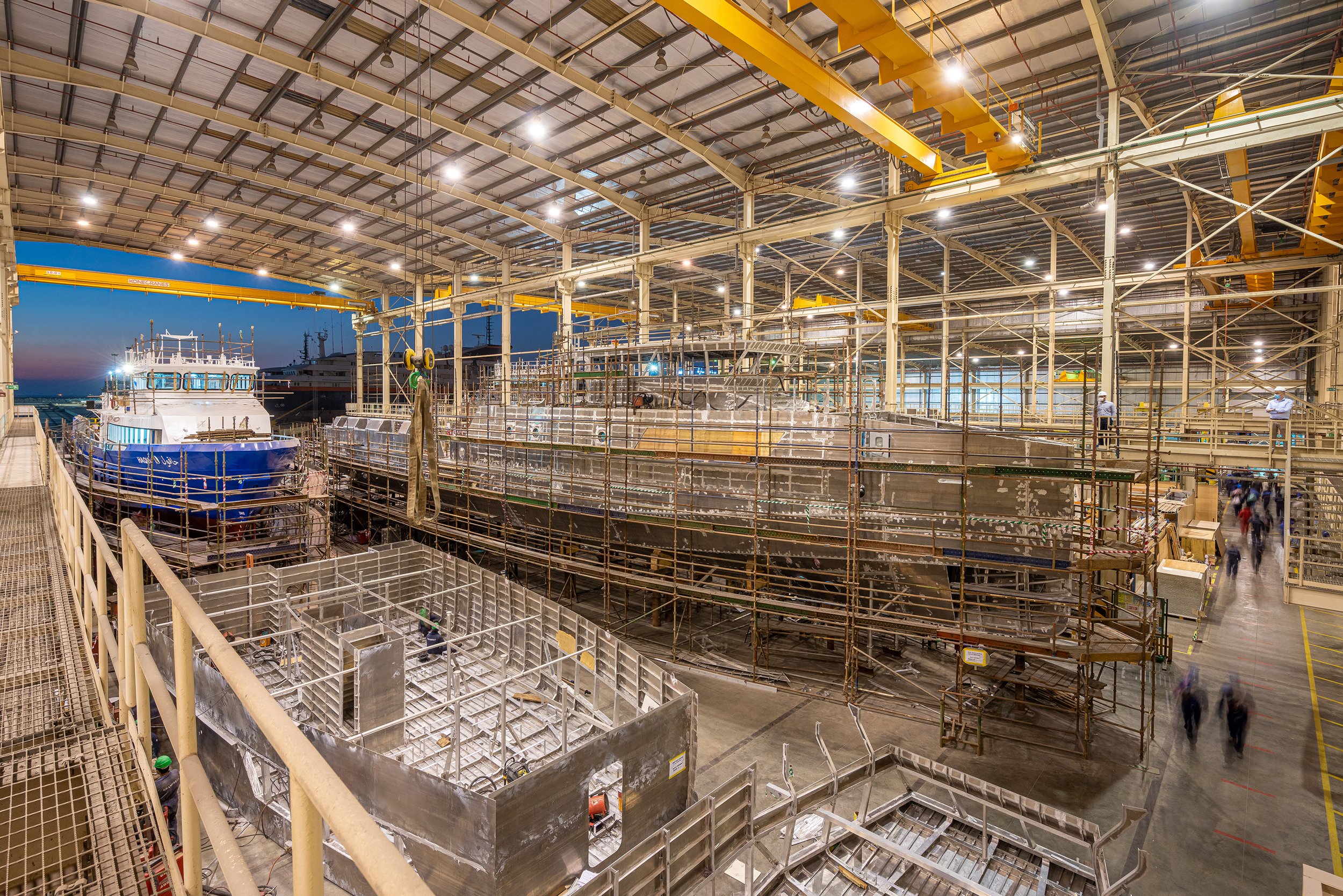 Interior of a shipyard or boat manufacturing facility with large boats in construction, scaffolding, and yellow overhead cranes.