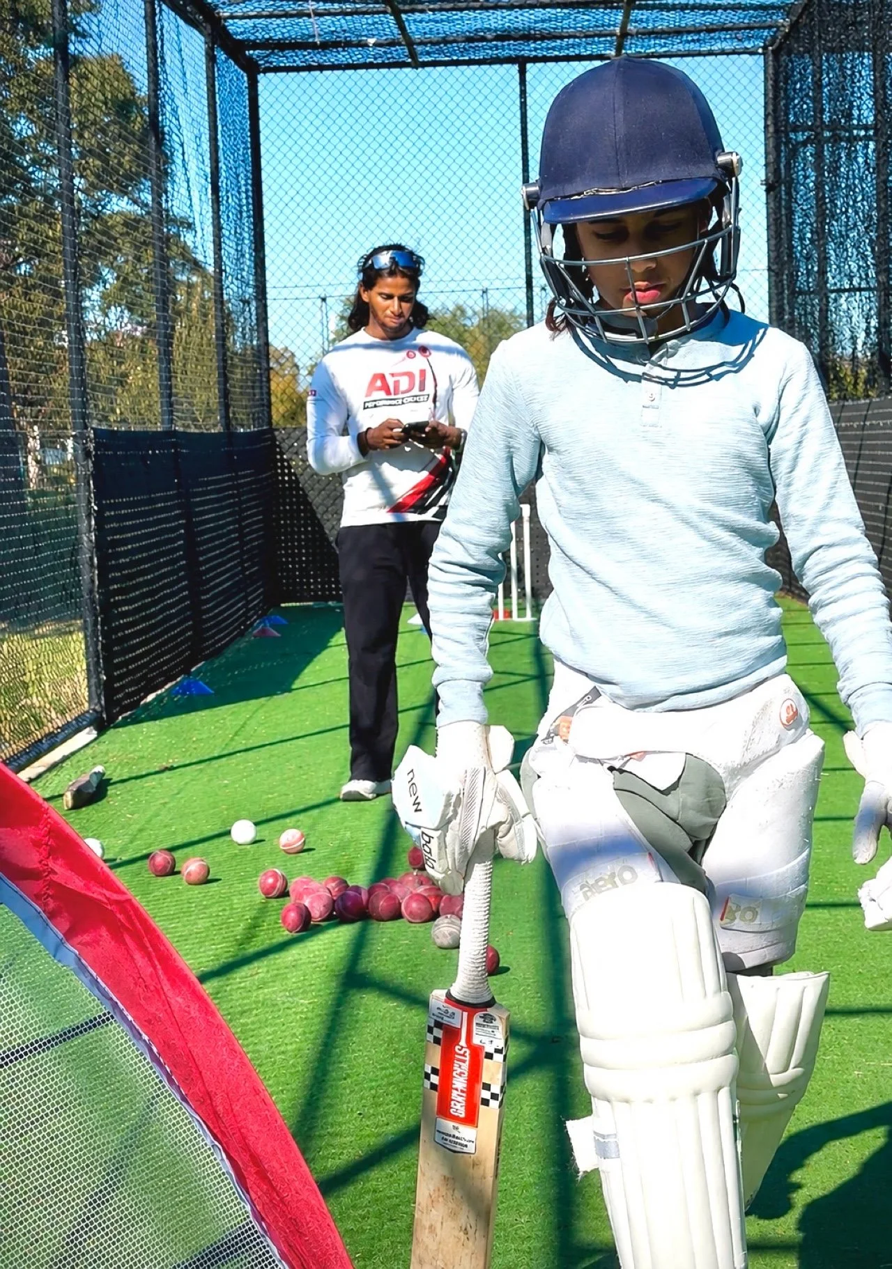 Cricket practice session with a young female wicketkeeper in gloves and pads, standing on a green artificial pitch, surrounded by scattered cricket balls, with another woman in the background using a smartphone inside a cricket net area