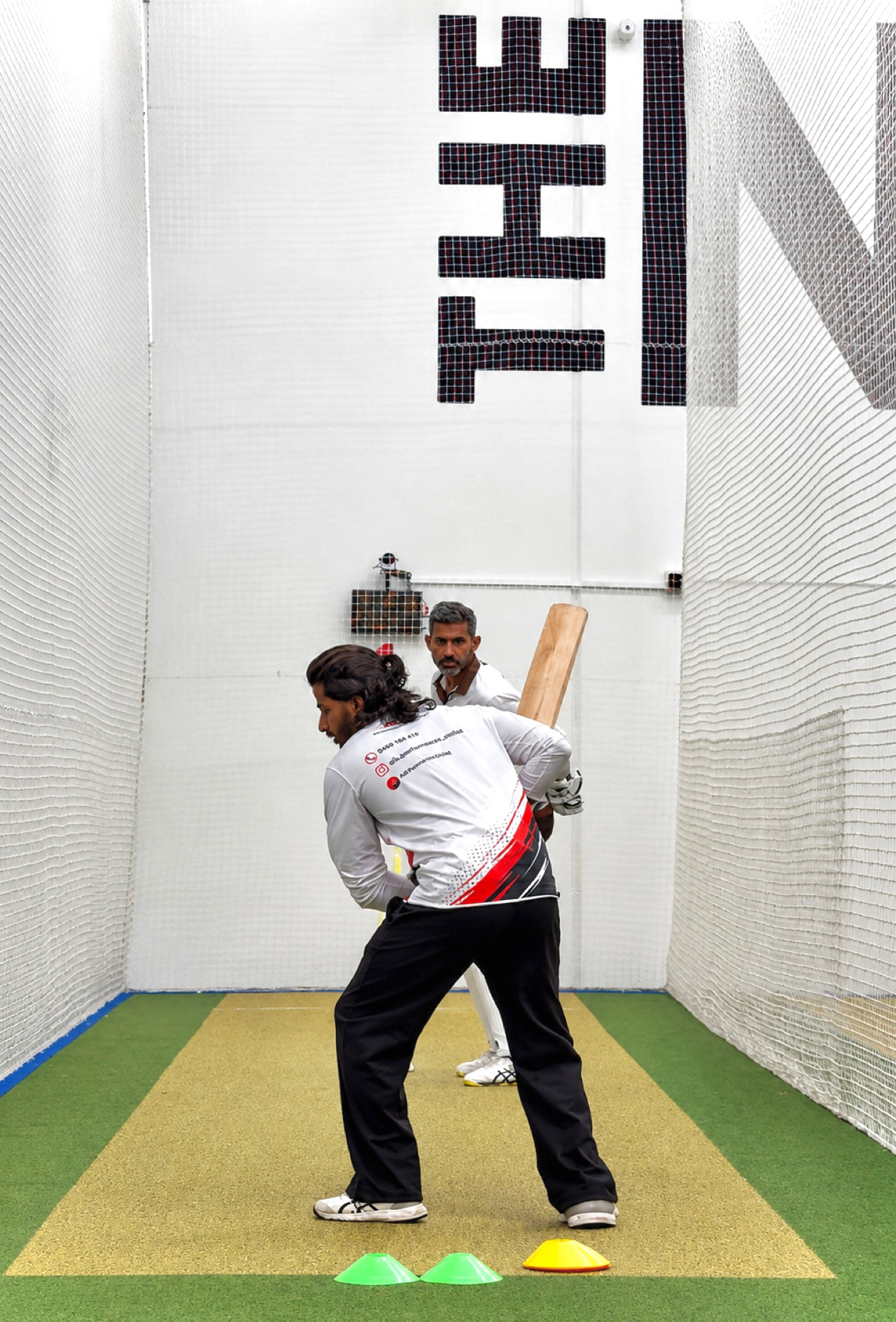 Two men practicing cricket bowling indoors, with cones set up on a yellow and green carpeted floor and a large sign in the background that reads 'HIT'.