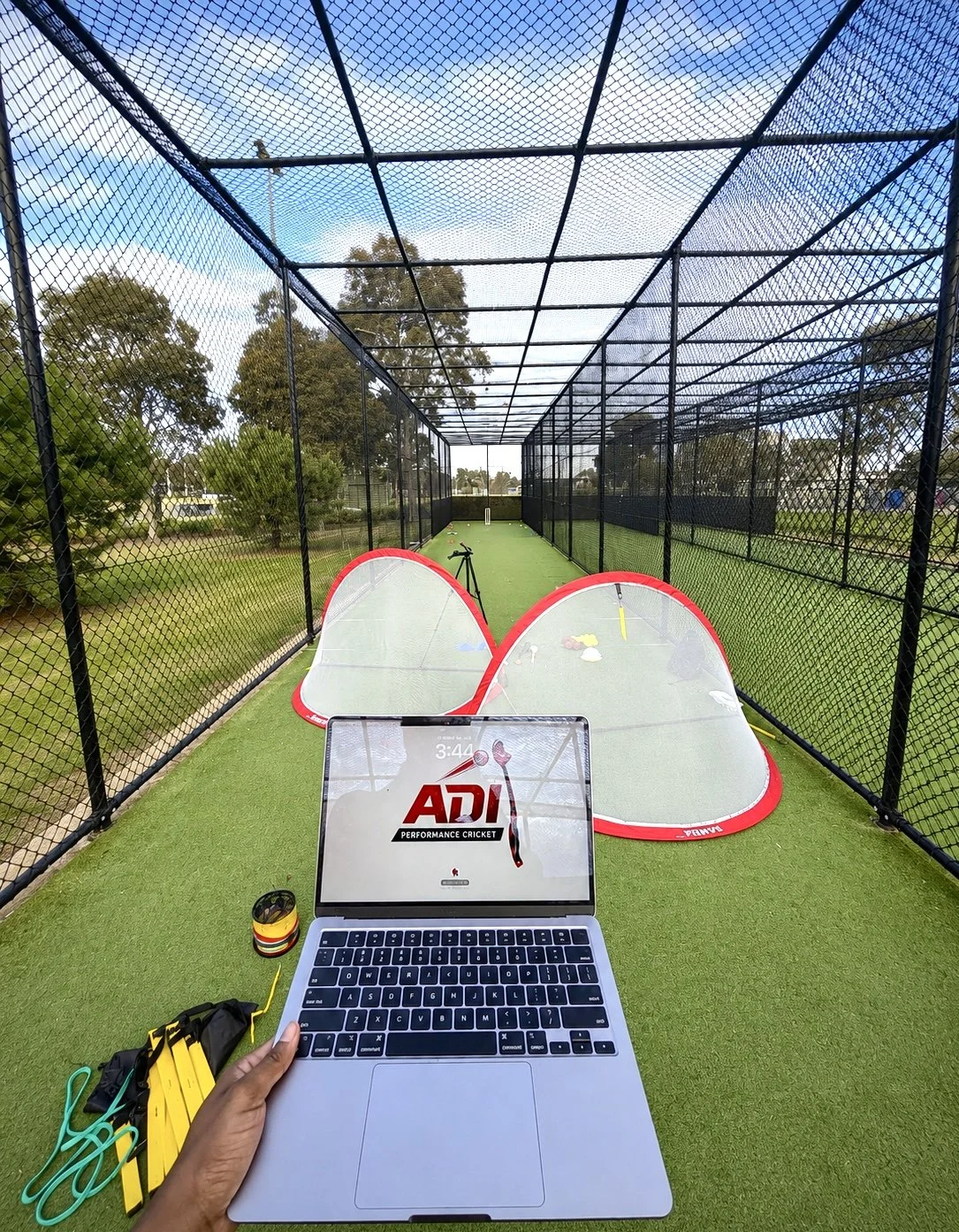 A cricket practice net with a laptop showing 'ADI Performance Cricket' on the screen, surrounded by cricket equipment including two pop-up wickets with a red border, a yellow cricket bat, and various training accessories, on a green turf with trees outside the netted enclosure.