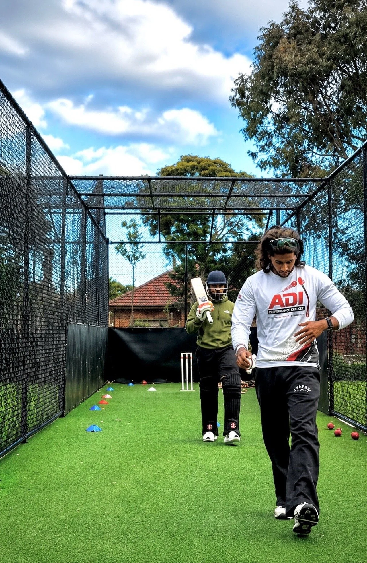 Cricket training session on an outdoor artificial pitch with two players practicing walking, one wearing a white shirt with 'ADI PERFORMANCE CRICKET' logo and the other in green, surrounded by various training cones and cricket equipment.