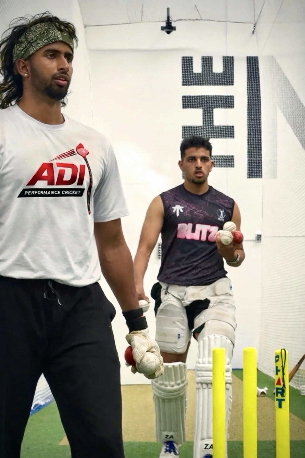 A cricket player in white protective gear holding a cricket ball stands next to a coach or instructor in a cricket training room, with cricket stumps and a batting net in the background.