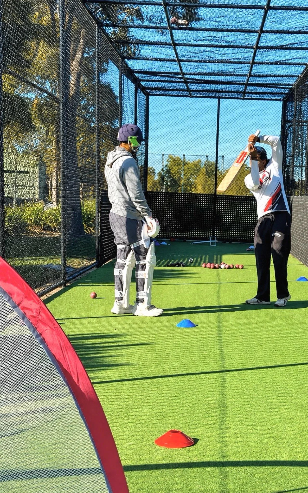 Two men in a cricket practice net, one dressed as a wicket keeper with pads and helmet, the other holding a cricket bat and wearing sunglasses, with cricket balls and cones on the green turf.