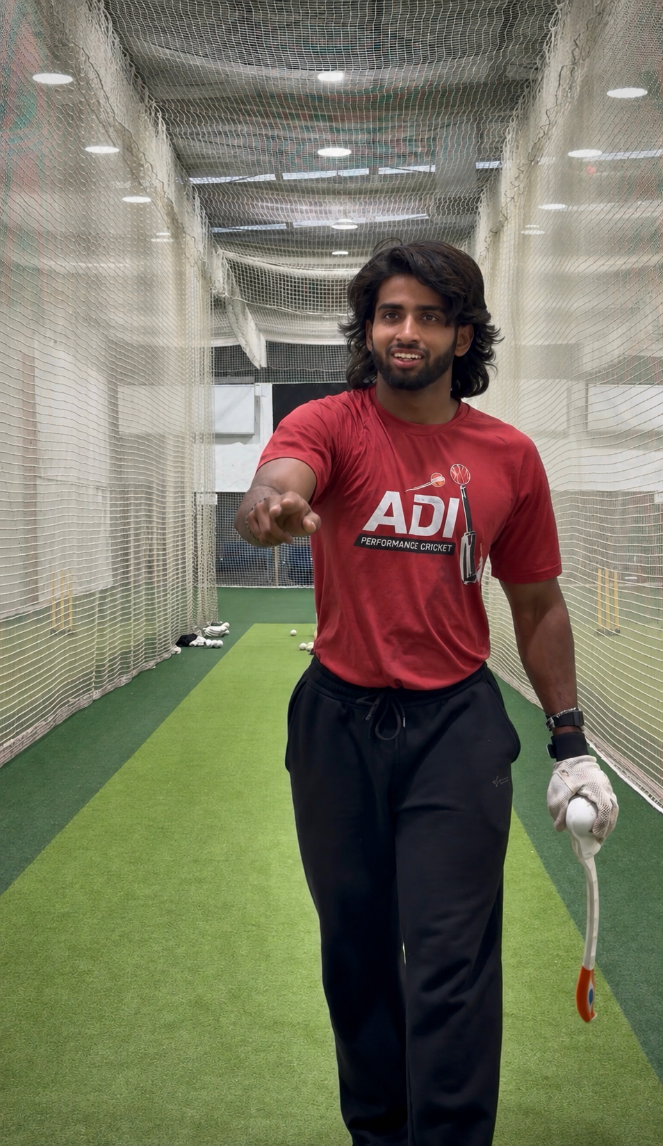 A man with shoulder-length dark hair and a beard, wearing a red ADI Performance Cricket t-shirt and black track pants, stands in an indoor cricket net. He is pointing forward with his right hand and holding a cricket bat in his left hand, which is protected with a white batting glove. The cricket ball is placed on the ground near him, with scattered cricket balls and shoes in the background.