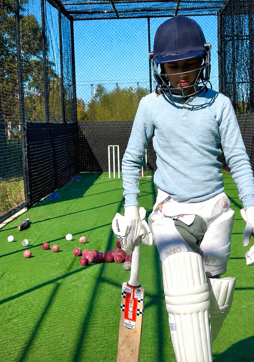 A young cricket player in batting gear, including a helmet and pads, on a practice pitch with scattered cricket balls and a cricket bat, within a netted outdoor cricket training area.