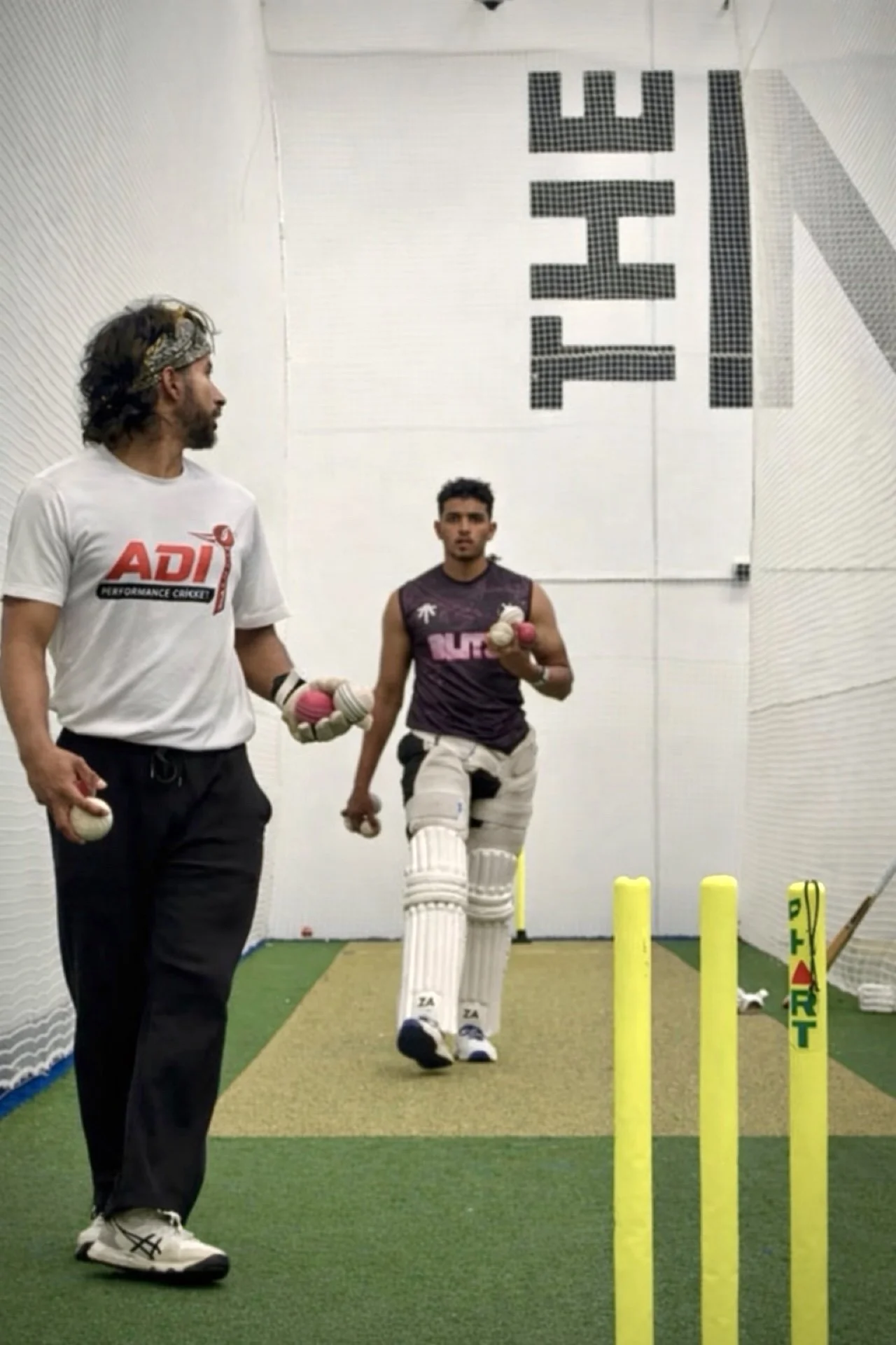 Cricket player practicing with coach in indoor cricket training facility, focusing on batting drills using training aids.