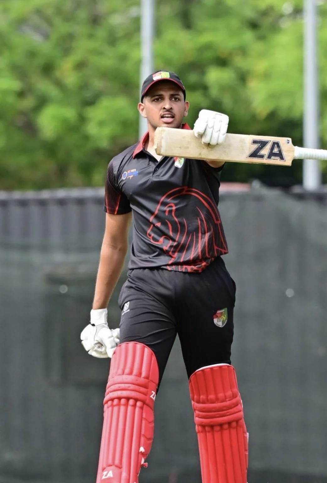 Cricket player holding a bat during practice, wearing a black and red uniform with a lion logo, red leg pads, and white gloves.