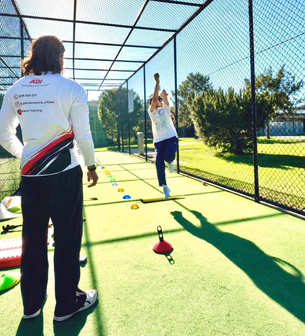 A person performs a cricket drill on a green practice mat inside a netted enclosure, while a coach observes. The person is mid-air, reaching for a ball, with cones and training equipment around.