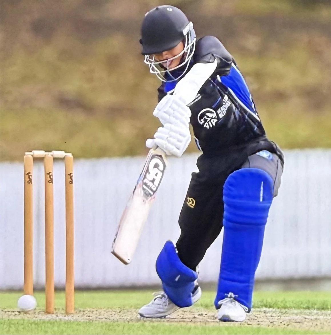 A young cricket player in full protective gear, including a helmet, pads, and gloves, attempts to hit a cricket ball with a cricket bat near the stumps on an outdoor field.