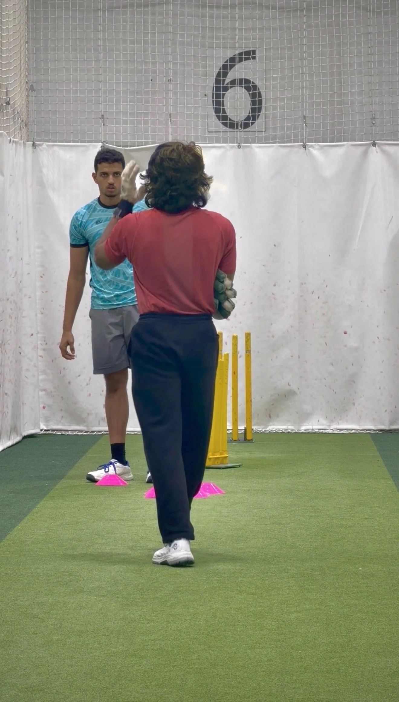 A coach instructs a young athlete in a sports training facility. The athlete is standing behind pink cones, and the coach is facing him, giving directions. Another person is in the background, standing near yellow training poles, with a wall marked by the number 6 above.