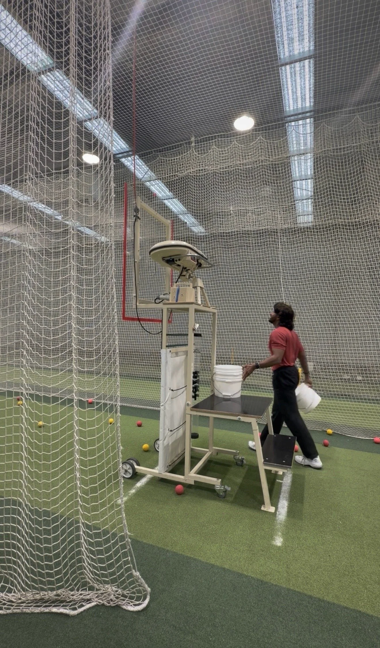 Indoor sports training area with netted walls, a person in athletic clothing holding a bucket, and a pitching machine on a rolling stand surrounded by scattered colored balls.