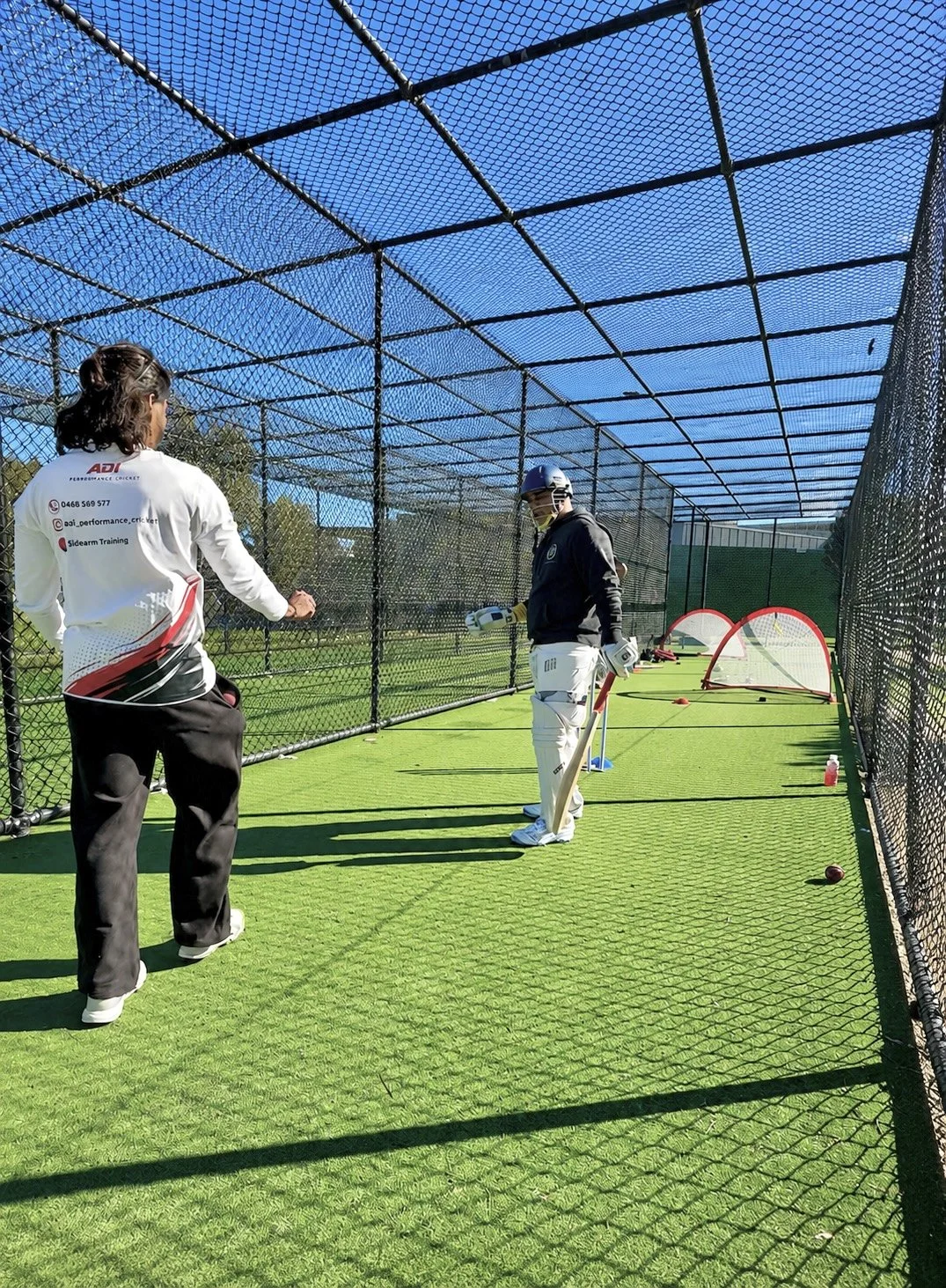 Cricketers on a practice pitch with training nets and mini goals, sunlight, clear blue sky.