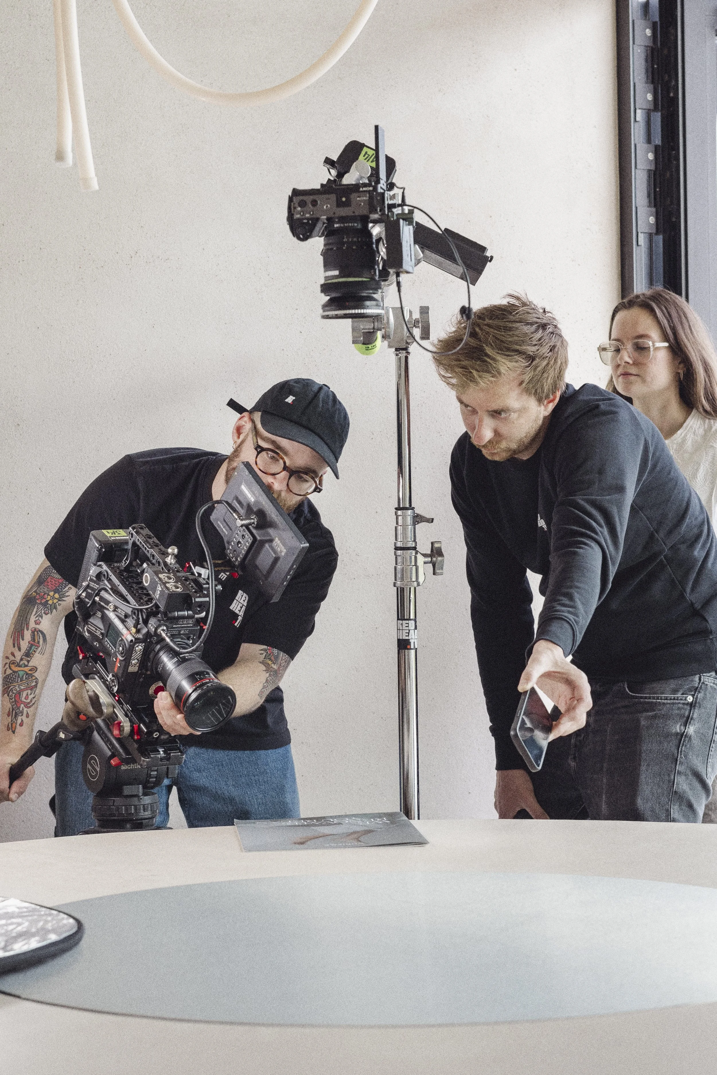 Three people are working together on a film or photography shoot indoors, with a camera mounted on a tripod and a large light stand.
