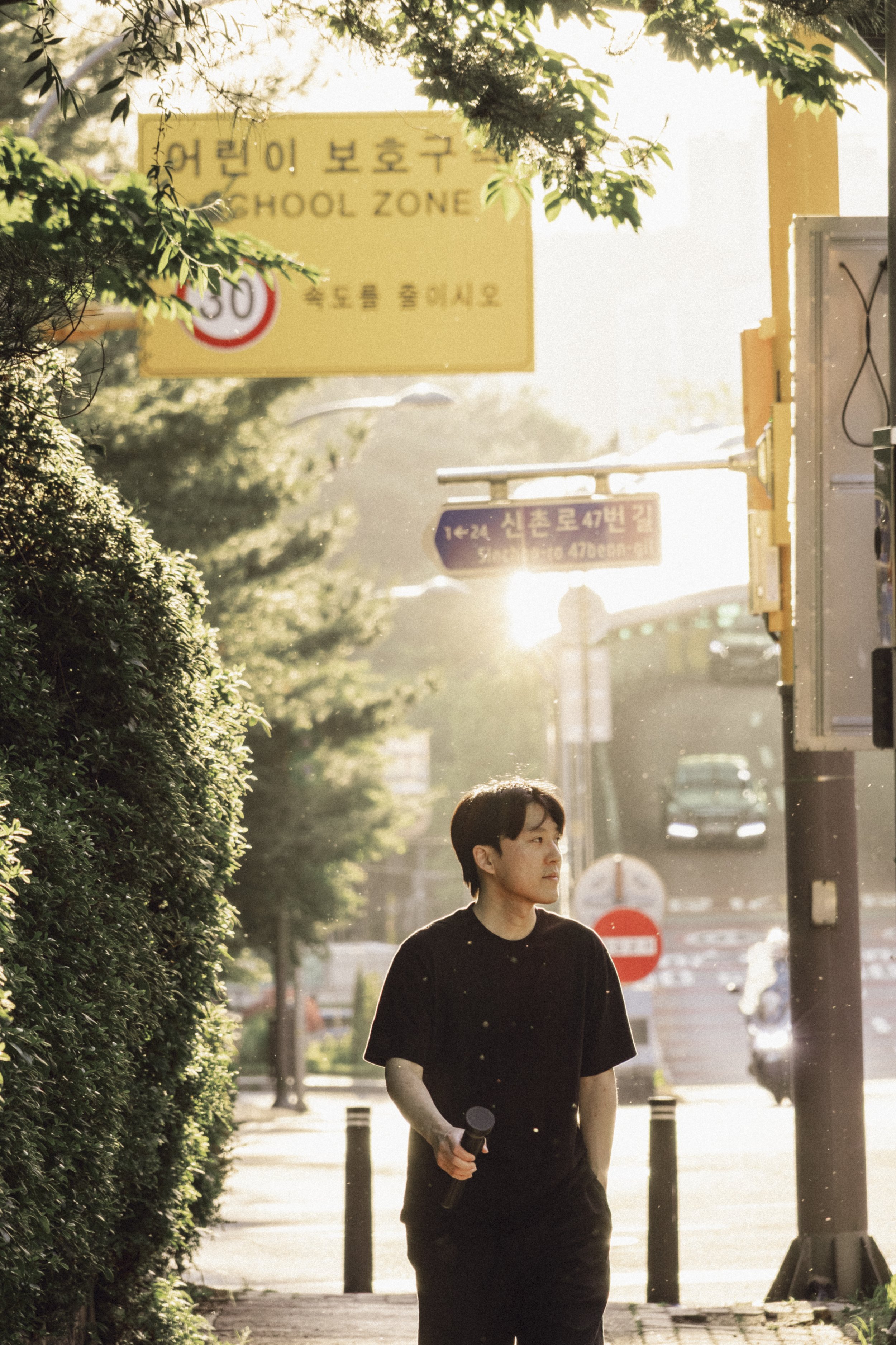A young man walking outdoors holding a tripod, with greenery and street signs in the background during sunset.