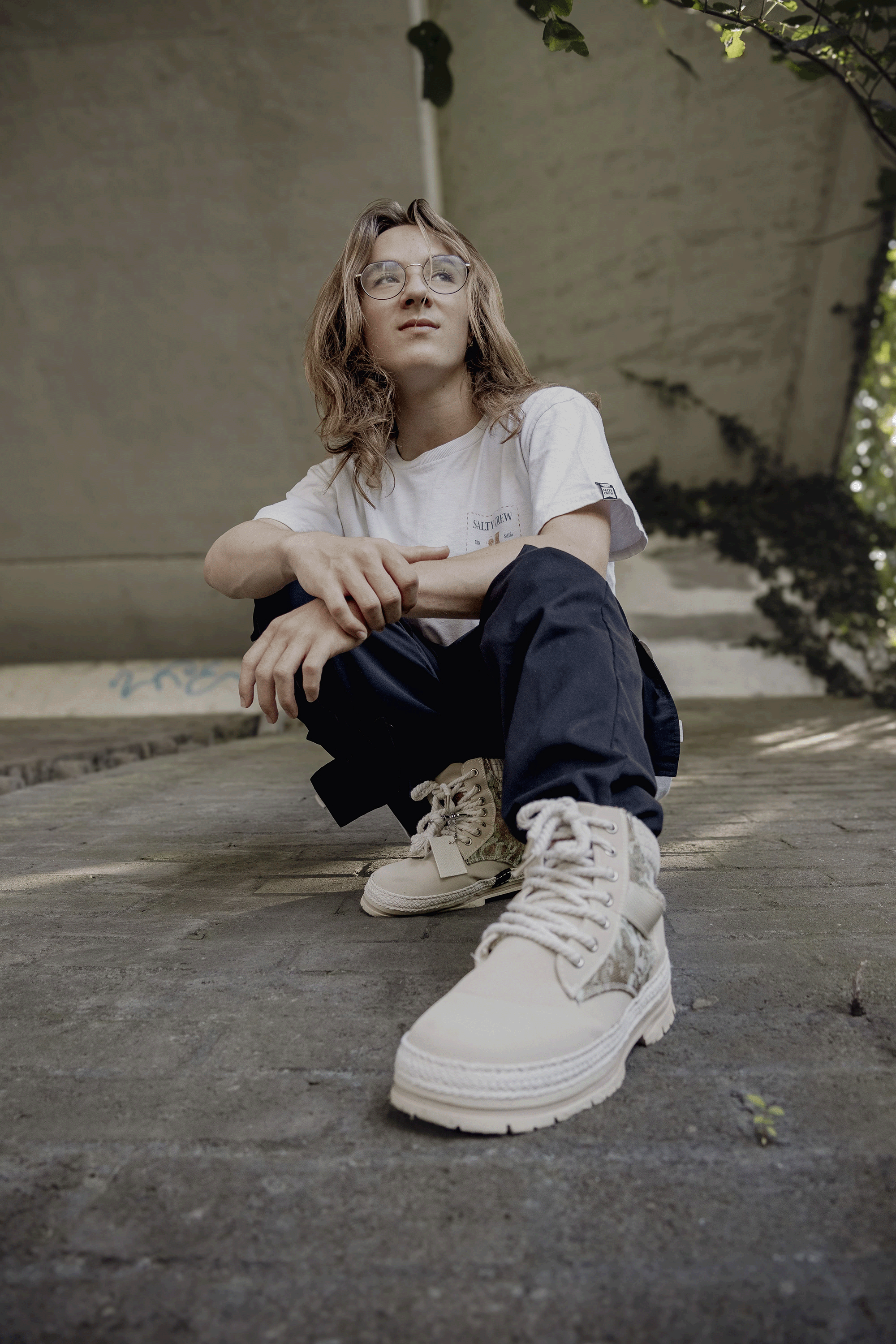 A young man with wavy light brown hair, wearing glasses, a white T-shirt, black pants, and beige sneakers, sitting on a concrete surface outdoors under a bridge.