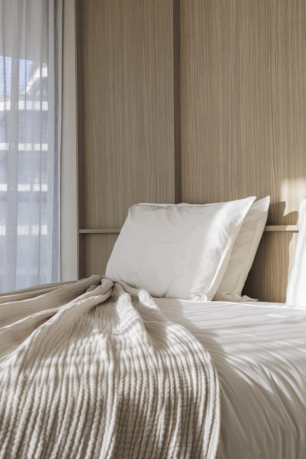 A neatly made bed with white pillows, beige blankets, and a wooden headboard in a bright room with a window and curtains.