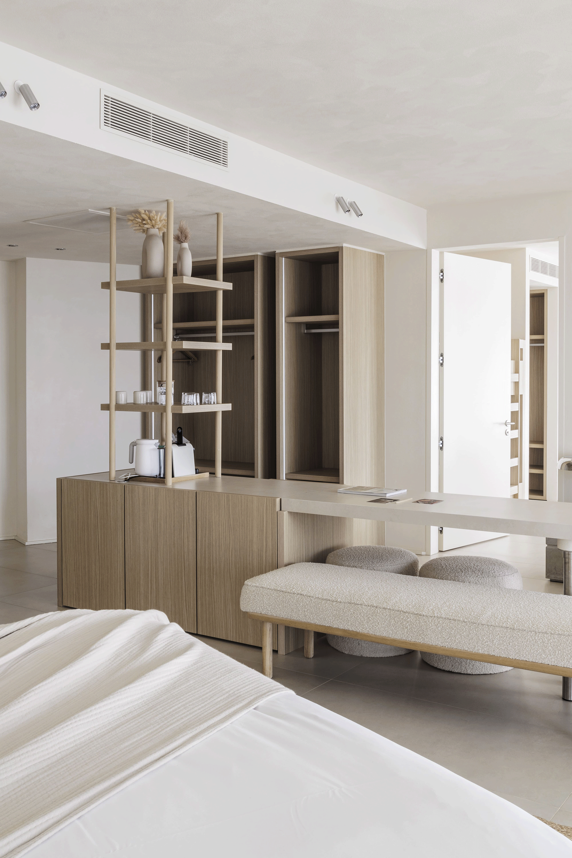 A modern bedroom with a bed, a beige upholstered bench, and wooden open closet shelving with decorative vases and glassware.