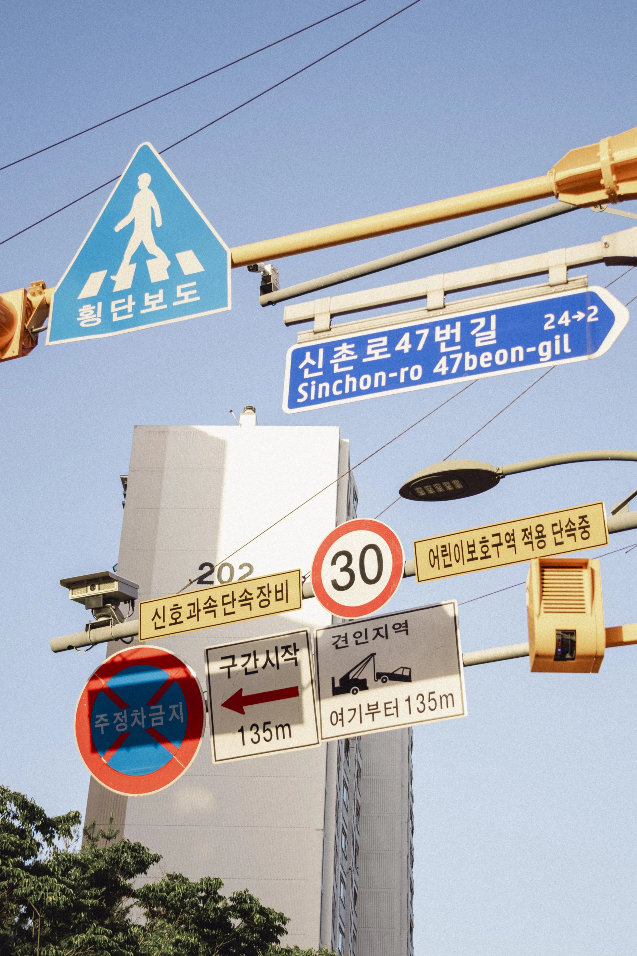 Street signs and signals in Korean on a city street. Blue pedestrian crossing sign, blue street name sign indicating 'Sinchon-ro 47beon-gil'. Several traffic and regulation signs, including speed limits and parking restrictions, with a tall building 