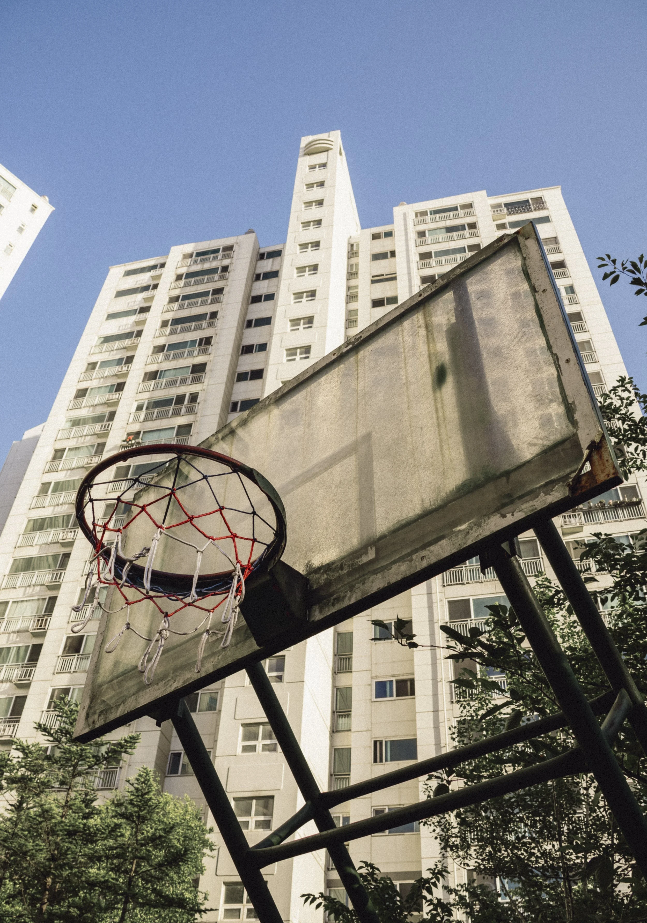 Low-angle view of a basketball hoop with a white backboard against a clear blue sky, surrounded by tall white apartment buildings and some green trees.
