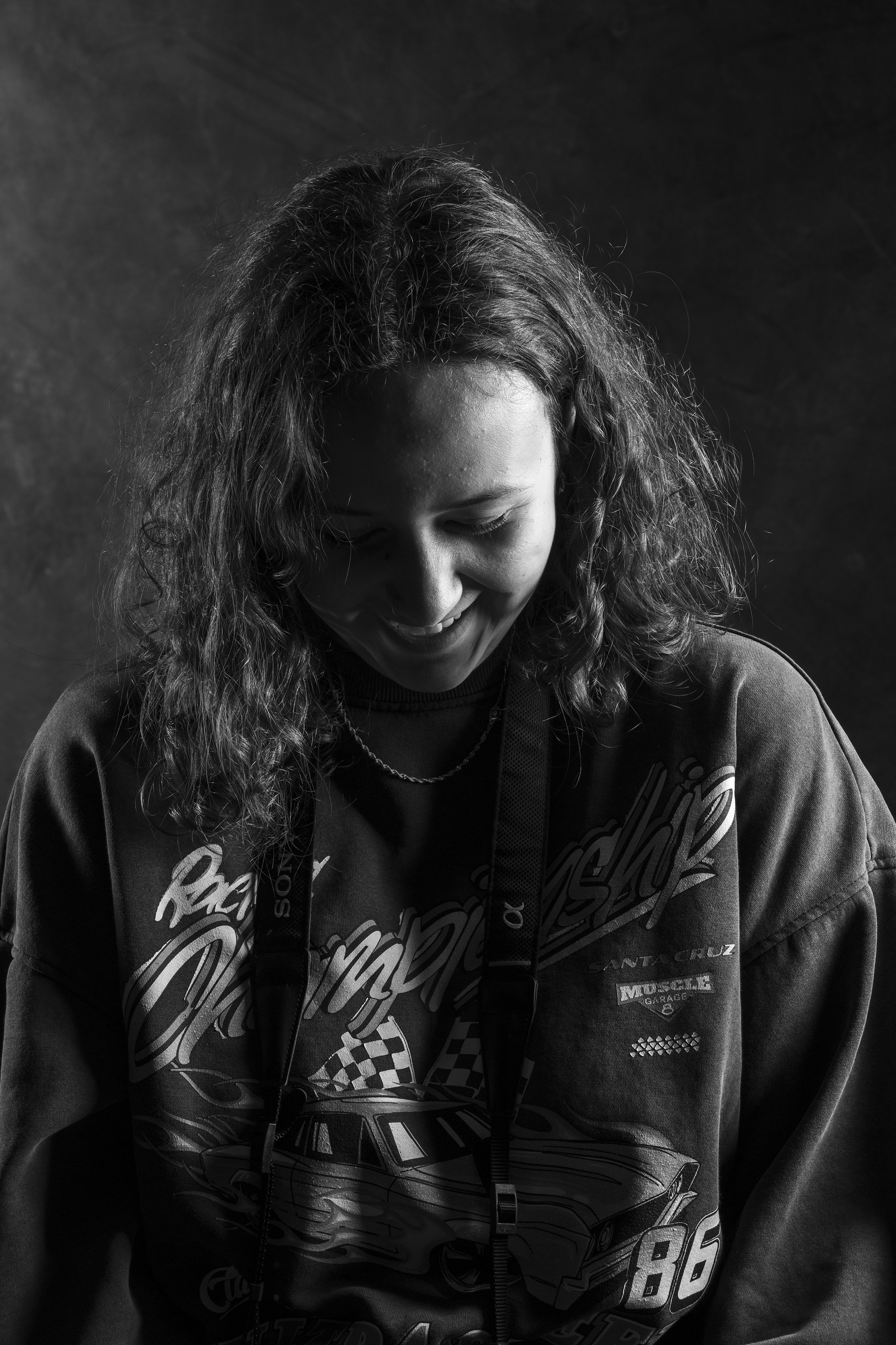 Black and white photo of a woman with curly hair, smiling and looking downward, wearing a Racing themed hoodie and a lanyard.