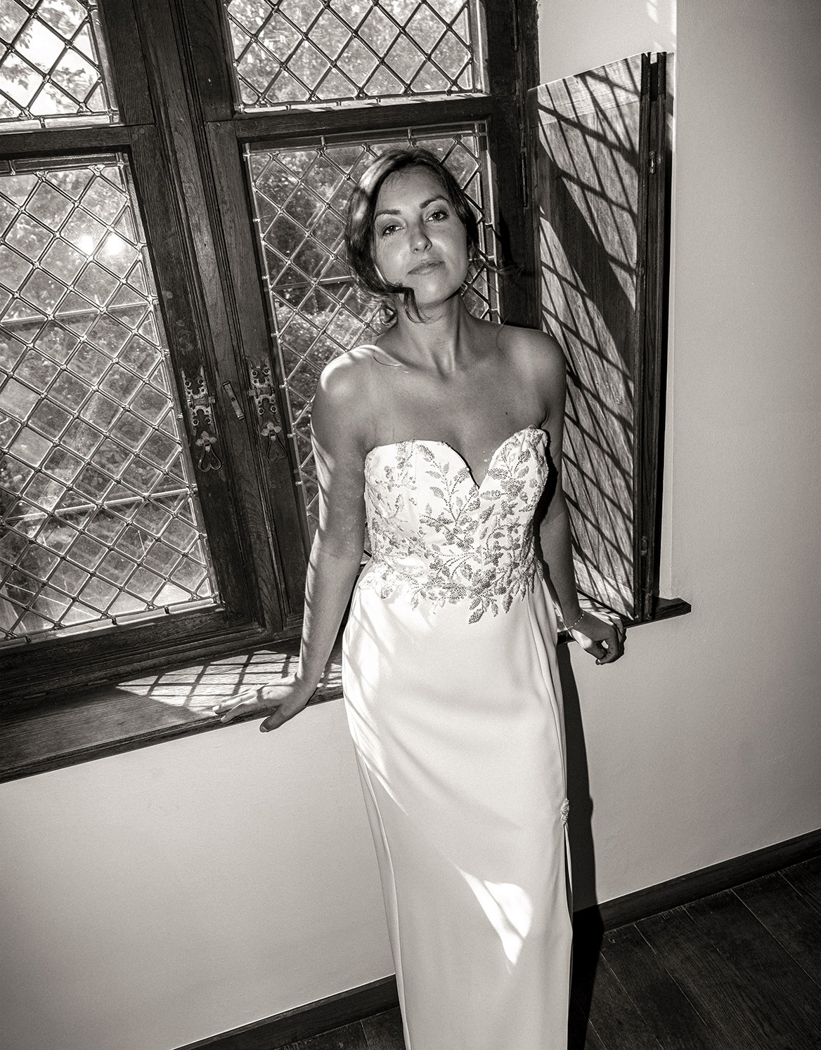 A woman in a strapless wedding dress with intricate embroidery, standing next to an open window with a diamond-patterned glass pane, casting shadows on her. She has short, wavy hair and a confident expression.