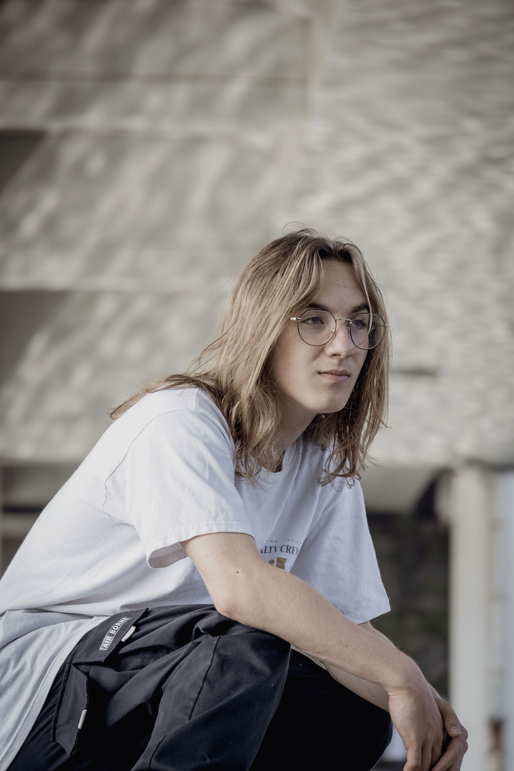 A young man with blonde hair, glasses, and a white t-shirt sitting outdoors.