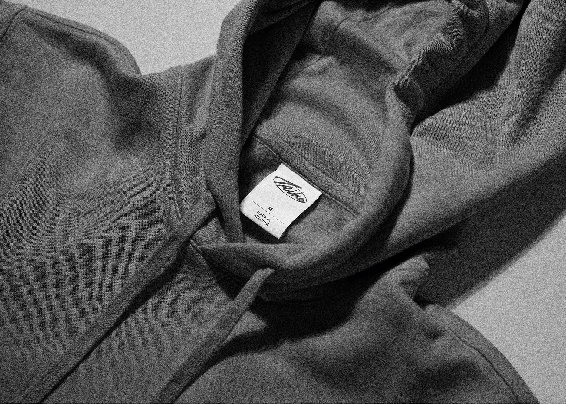 Close-up of a dark-colored hoodie with the hood slightly open, revealing a white clothing label that says 'Triko' and 'Made in Belgium'.
