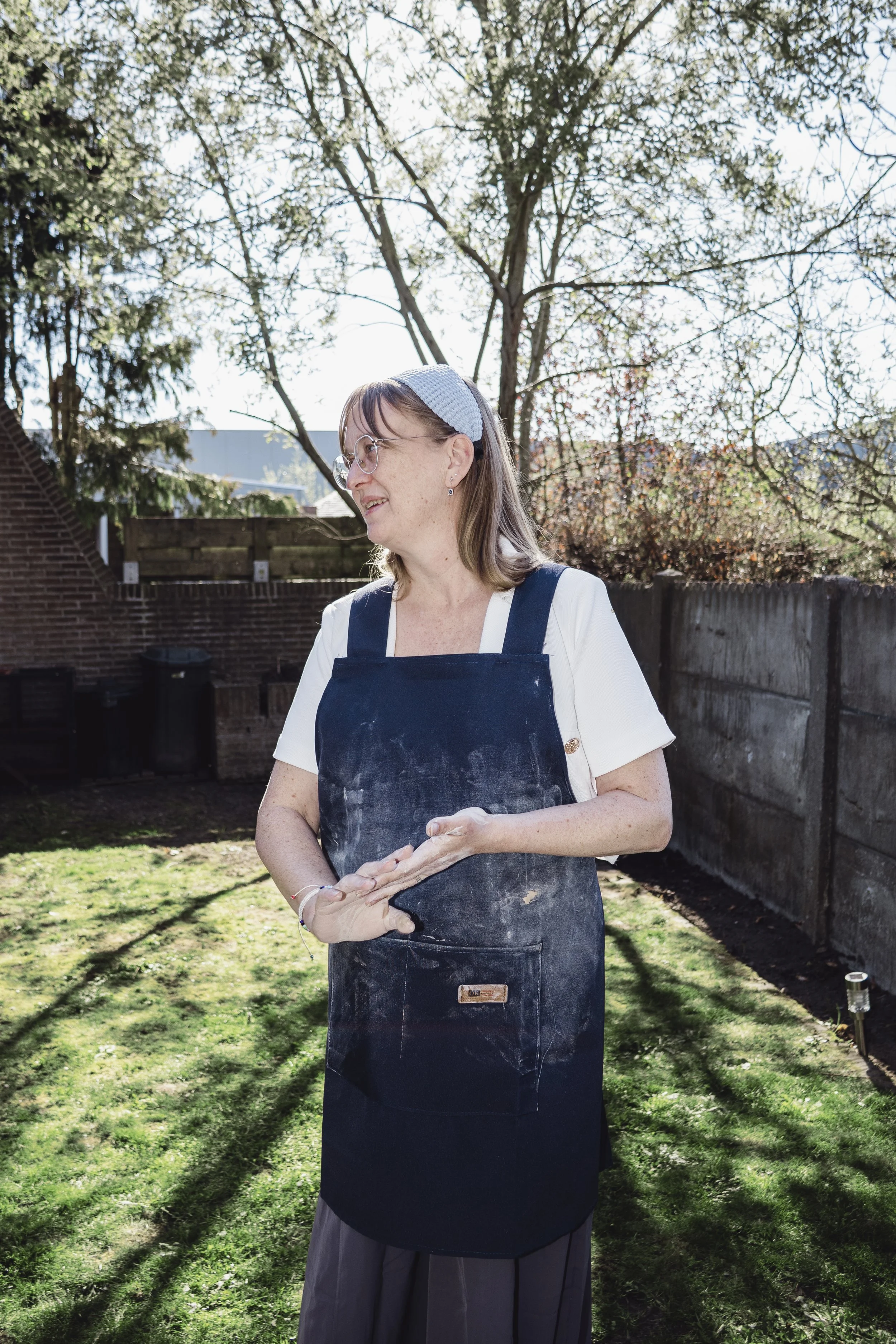 A woman with glasses and a blue headband, wearing a white shirt and a dark apron, standing outdoors on a sunny day with trees and a wooden fence in the background.