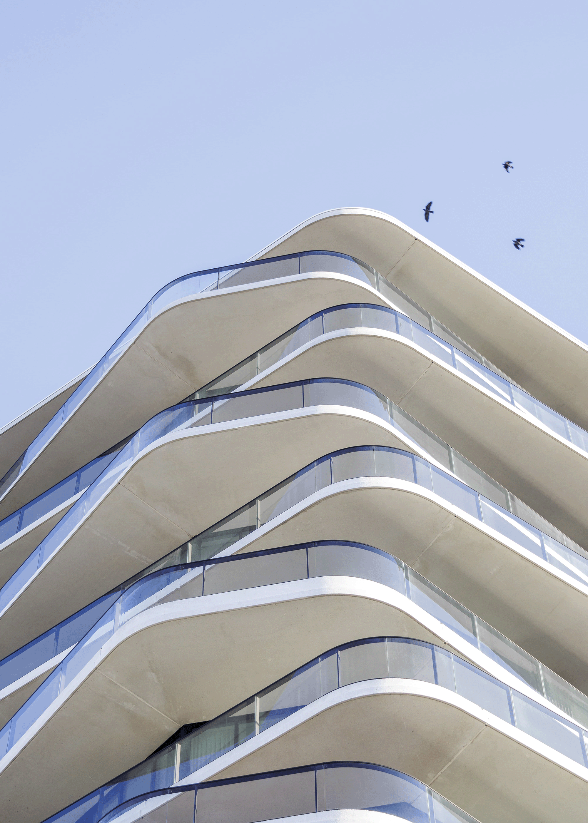 Modern white building with rounded balconies and glass railings, viewed from below, clear blue sky, birds flying overhead.
