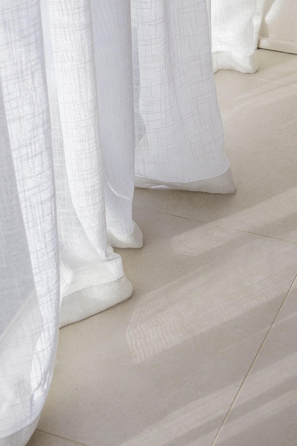 Close-up of white sheer curtains and sunlight casting shadows on beige tiled floor.
