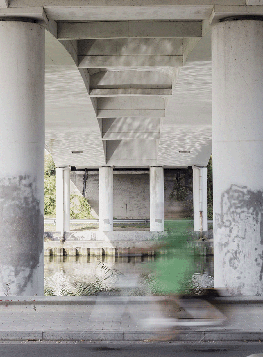 Underneath a bridge with large concrete support columns, view of water and a blurred cyclist passing by underneath.