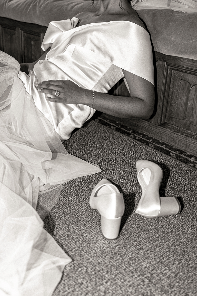 Person lying on the floor next to a bed, wearing a satin dress, with white high-heeled shoes removed nearby.